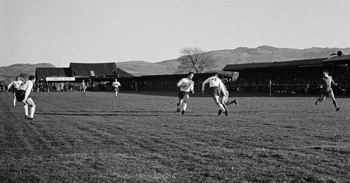 Fotografía antigua en blanco y negro de una jugada de ataque con la tribuna y las montañas al fondo